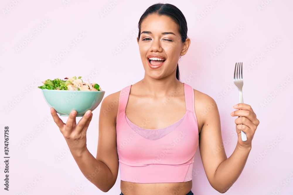 Young beautiful latin girl wearing sportswear holding salad winking looking at the camera with sexy expression, cheerful and happy face. Young beautiful latin girl wearing sportswear holding salad winking looking at the camera with sexy expression, cheerful and happy face.