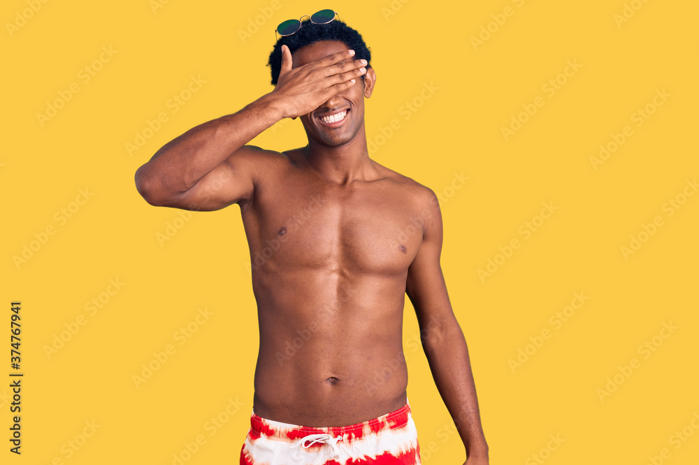African handsome man wearing swimsuit and sunglasses smiling and laughing with hand on face covering eyes for surprise. blind concept.