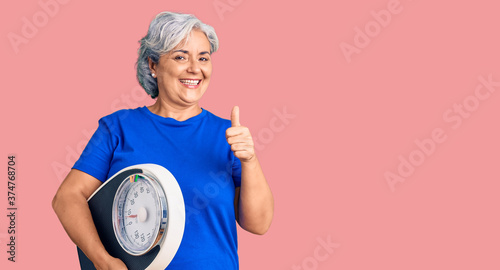 Senior woman with gray hair holding weight machine to balance weight loss smiling happy and positive, thumb up doing excellent and approval sign © Krakenimages.com