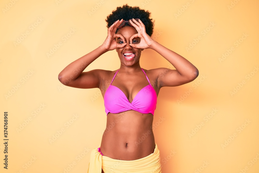 Young african american woman wearing bikini doing ok gesture like binoculars sticking tongue out, eyes looking through fingers. crazy expression. Young african american woman wearing bikini doing ok gesture like binoculars sticking tongue out, eyes looking through fingers. crazy expression.