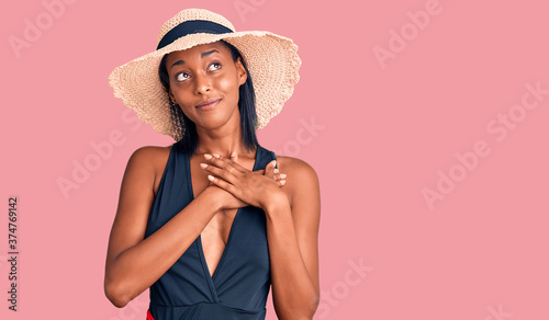 Young african american woman wearing swimsuit and summer hat smiling with hands on chest with closed eyes and grateful gesture on face. health concept.