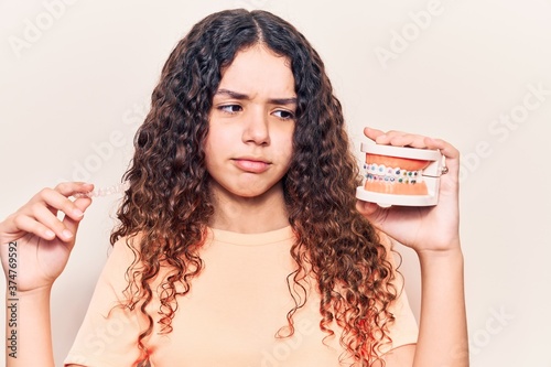 Adorable latin teenager holding denture with bracket and aligner standing over isolated white background