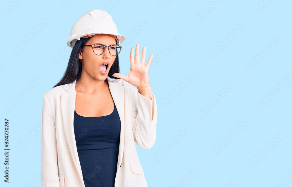 Young beautiful latin girl wearing architect hardhat and glasses shouting and screaming loud to side with hand on mouth. communication concept. Young beautiful latin girl wearing architect hardhat and glasses shouting and screaming loud to side with hand on mouth. communication concept.