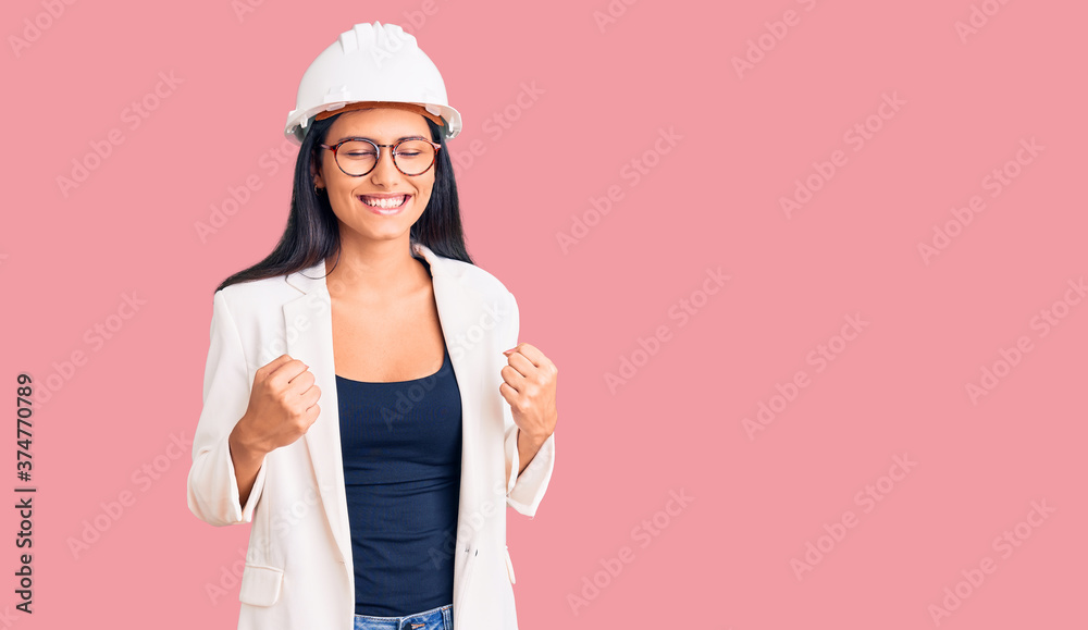 Young beautiful latin girl wearing architect hardhat and glasses excited for success with arms raised and eyes closed celebrating victory smiling. winner concept. Young beautiful latin girl wearing architect hardhat and glasses excited for success with arms raised and eyes closed celebrating victory smiling. winner concept.