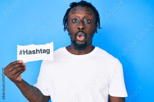 Young african american man with braids holding hashtag paper scared and amazed with open mouth for surprise, disbelief face © Krakenimages.com