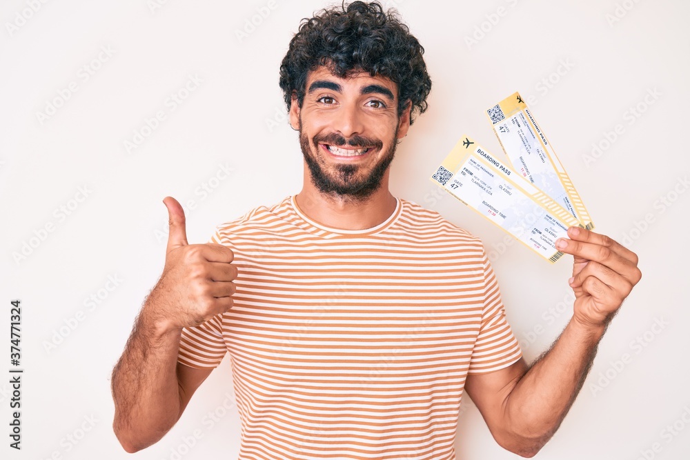 Handsome young man with curly hair and bear holding boarding pass smiling happy and positive, thumb up doing excellent and approval sign Handsome young man with curly hair and bear holding boarding pass smiling happy and positive, thumb up doing excellent and approval sign