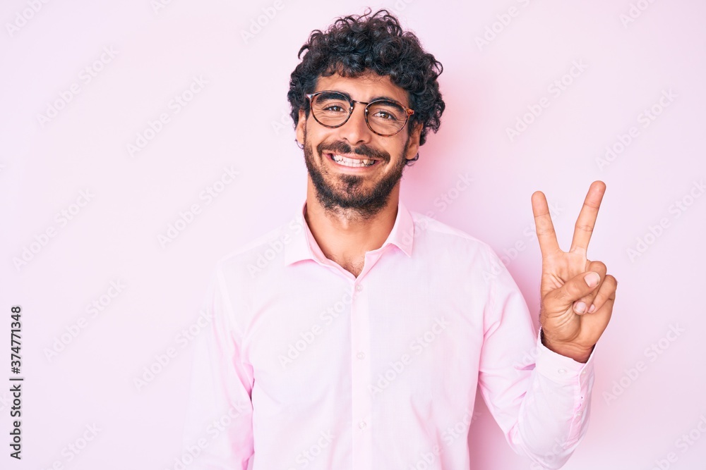 Handsome young man with curly hair and bear wearing business shirt and glasses smiling with happy face winking at the camera doing victory sign. number two.