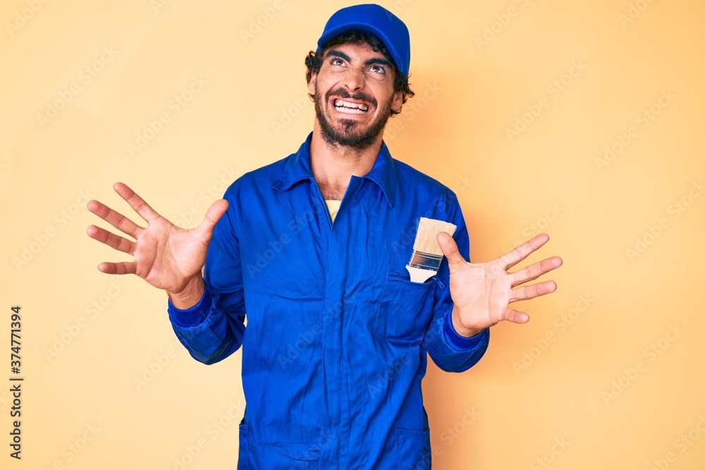 Handsome young man with curly hair and bear wearing builder jumpsuit uniform crazy and mad shouting and yelling with aggressive expression and arms raised. frustration concept.