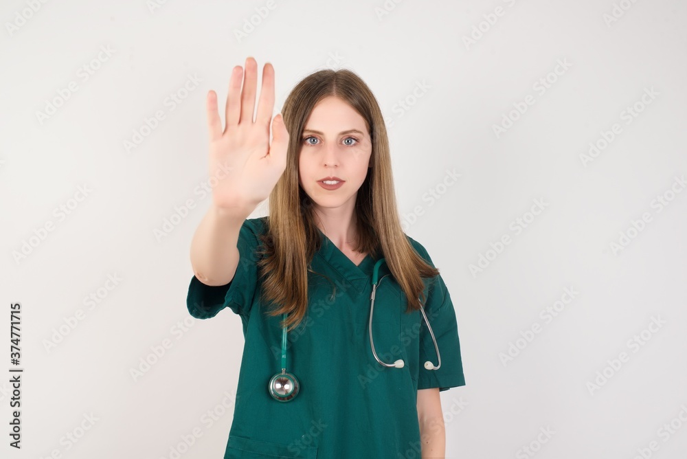 Female doctor wearing a green scrubs and stethoscope doing stop gesture with palm of the hand. Warning expression with negative and serious gesture on the face Female doctor wearing a green scrubs and stethoscope doing stop gesture with palm of the hand. Warning expression with negative and serious gesture on the face
