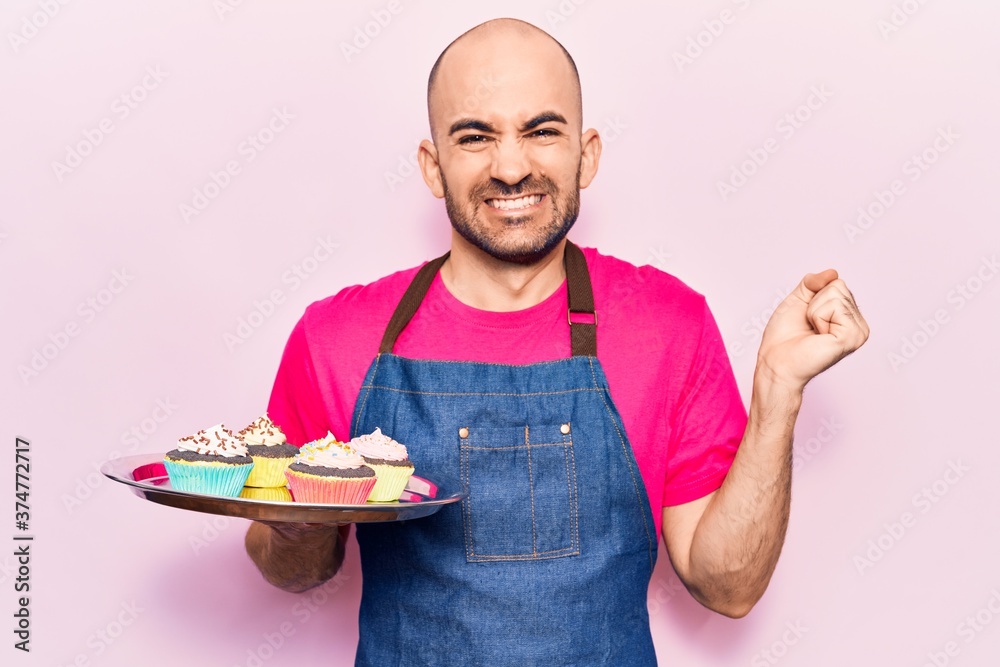 Young handsome bald man wearing apron holding cupcakes screaming proud, celebrating victory and success very excited with raised arm Young handsome bald man wearing apron holding cupcakes screaming proud, celebrating victory and success very excited with raised arm