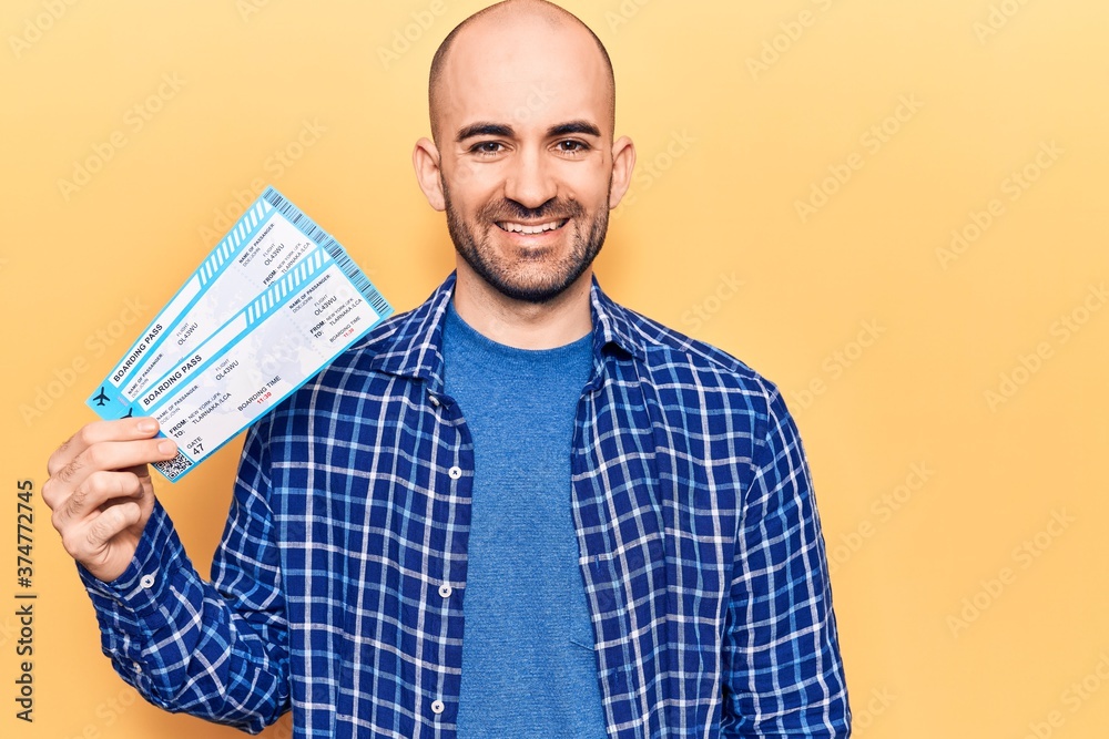 Young handsome bald man holding airline boarding pass looking positive and happy standing and smiling with a confident smile showing teeth Young handsome bald man holding airline boarding pass looking positive and happy standing and smiling with a confident smile showing teeth