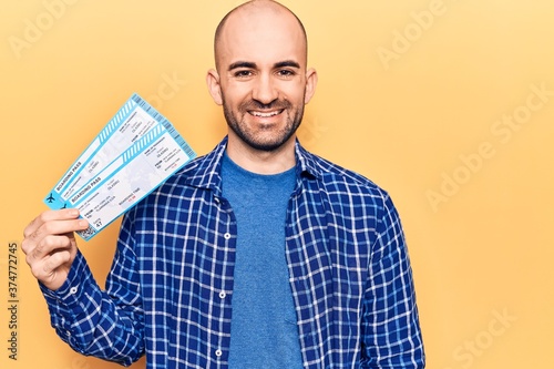 Young handsome bald man holding airline boarding pass looking positive and happy standing and smiling with a confident smile showing teeth photo