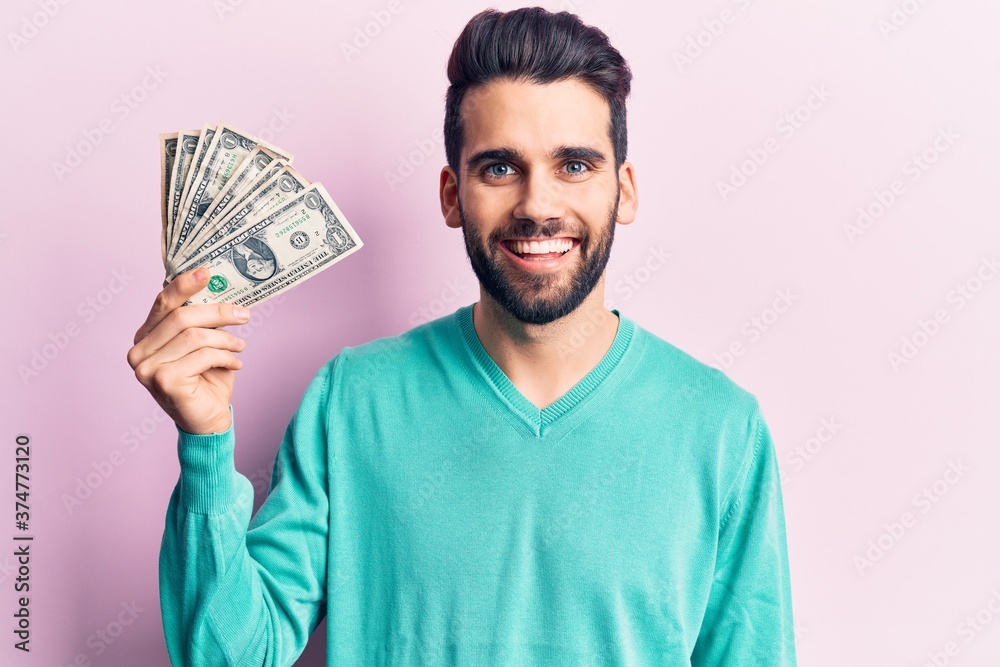 Young handsome man with beard holding bunch of dollar looking positive and happy standing and smiling with a confident smile showing teeth Young handsome man with beard holding bunch of dollar looking positive and happy standing and smiling with a confident smile showing teeth