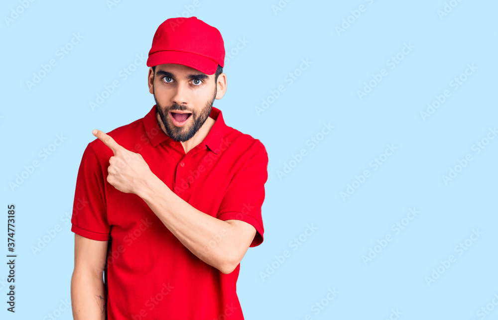 Young handsome man with beard wearing delivery uniform surprised pointing with finger to the side, open mouth amazed expression.