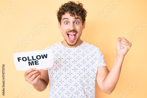 Young caucasian man with curly hair holding follow me message paper screaming proud, celebrating victory and success very excited with raised arms © Krakenimages.com