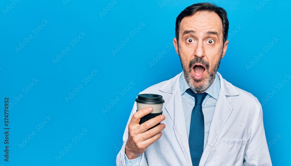 Middle age handsome doctor man wearing coat drinking cup of takeaway coffee scared and amazed with open mouth for surprise, disbelief face Middle age handsome doctor man wearing coat drinking cup of takeaway coffee scared and amazed with open mouth for surprise, disbelief face