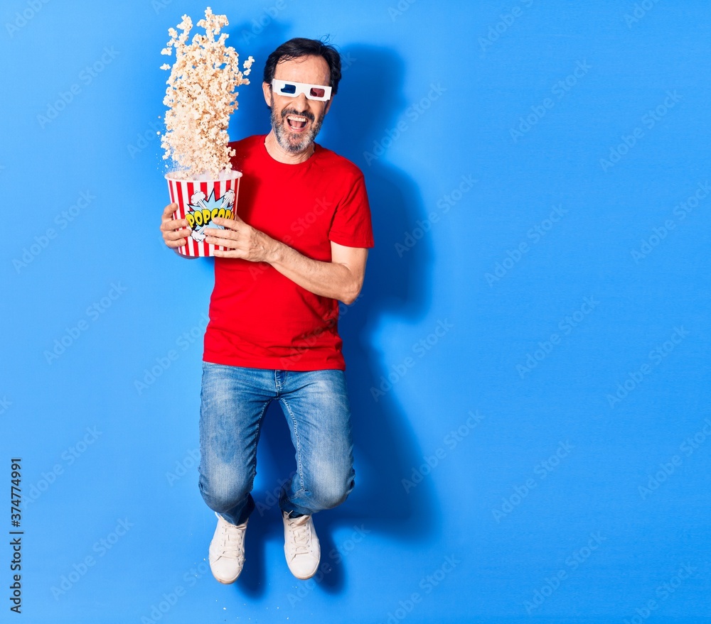 Middle age handsome hispanic man watching movie using 3d glasses similing happy. Holding bucket of popcorn jumping with smile on face over isolated blue background Middle age handsome hispanic man watching movie using 3d glasses similing happy. Holding bucket of popcorn jumping with smile on face over isolated blue background