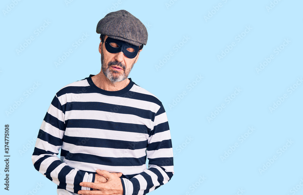 Middle age handsome man wearing burglar mask with hand on stomach because indigestion, painful illness feeling unwell. ache concept. Middle age handsome man wearing burglar mask with hand on stomach because indigestion, painful illness feeling unwell. ache concept.