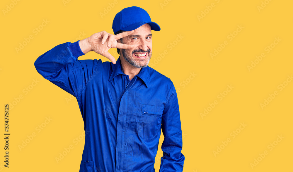 Middle age handsome man wearing mechanic uniform doing peace symbol with fingers over face, smiling cheerful showing victory