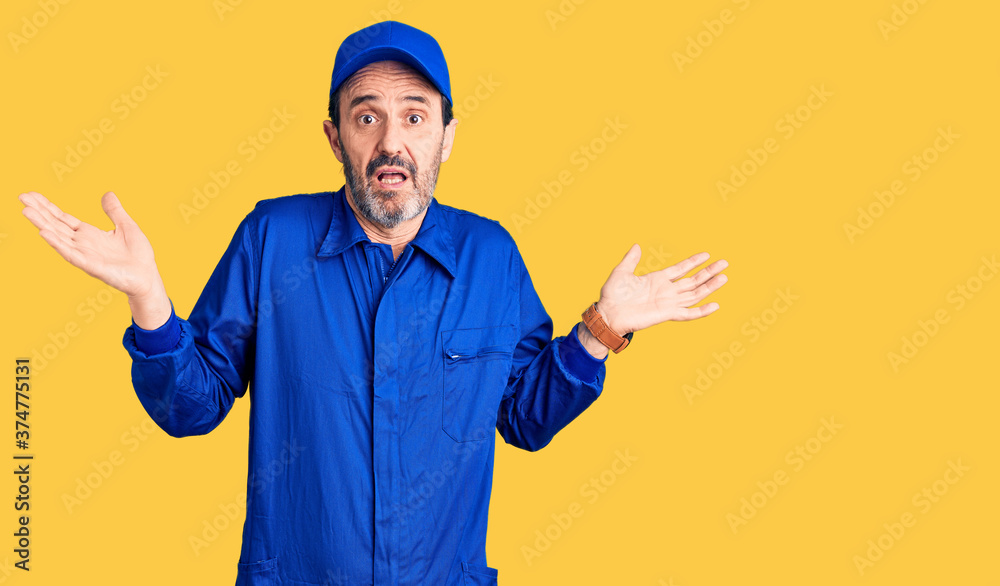Middle age handsome man wearing mechanic uniform clueless and confused expression with arms and hands raised. doubt concept.