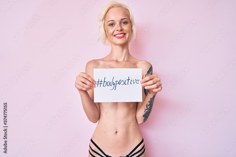 Young blonde woman with tattoo standing shirtless holding banner with body positive message smiling with a happy and cool smile on face. showing teeth. Young blonde woman with tattoo standing shirtless holding banner with body positive message smiling with a happy and cool smile on face. showing teeth.