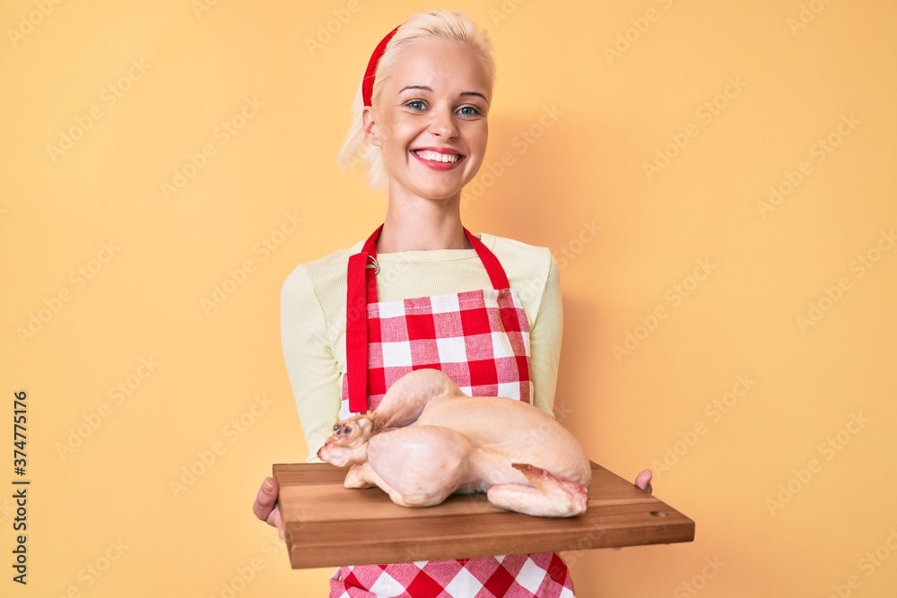 Young blonde woman wearing apron cooking chicken smiling with a happy and cool smile on face. showing teeth. Young blonde woman wearing apron cooking chicken smiling with a happy and cool smile on face. showing teeth.