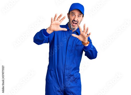 Middle age handsome man wearing mechanic uniform afraid and terrified with fear expression stop gesture with hands, shouting in shock. panic concept.