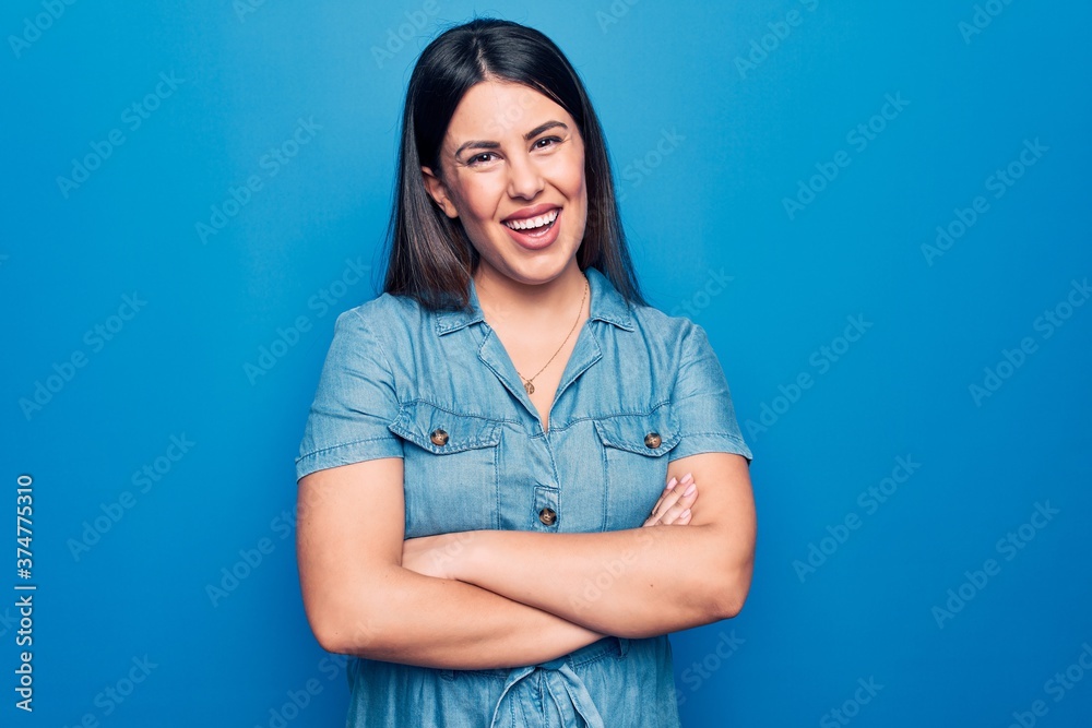 Young beautiful brunette woman wearing casual denim dress over isolated blue background happy face smiling with crossed arms looking at the camera. Positive person. Young beautiful brunette woman wearing casual denim dress over isolated blue background happy face smiling with crossed arms looking at the camera. Positive person.