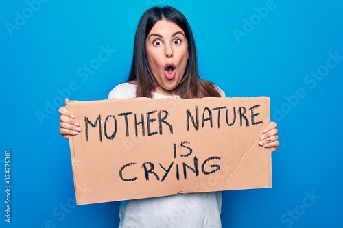 Young woman asking for environment care holding banner with mother nature is crying message scared and amazed with open mouth for surprise, disbelief face