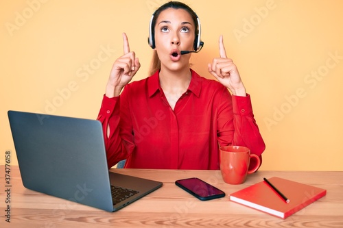 Young brunette woman wearing operator headset at the call center office amazed and surprised looking up and pointing with fingers and raised arms. photo