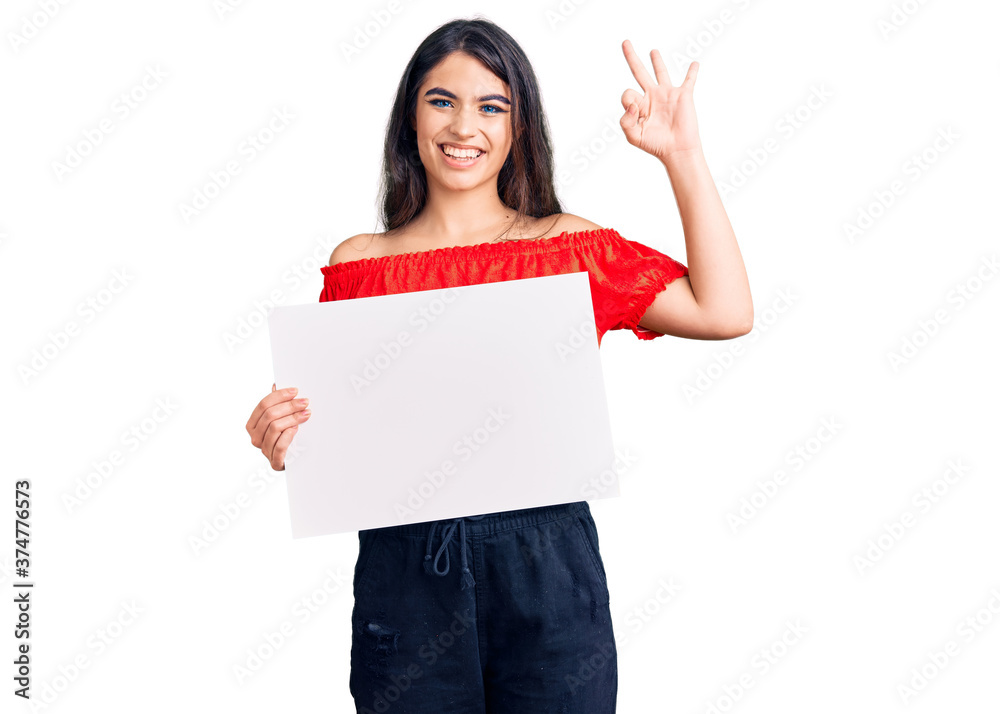Brunette teenager girl holding blank empty banner doing ok sign with fingers, smiling friendly gesturing excellent symbol Brunette teenager girl holding blank empty banner doing ok sign with fingers, smiling friendly gesturing excellent symbol