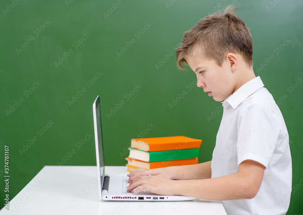 A boy is studying at a laptop while sitting at a school desk on the background of a green school board A boy is studying at a laptop while sitting at a school desk on the background of a green school board