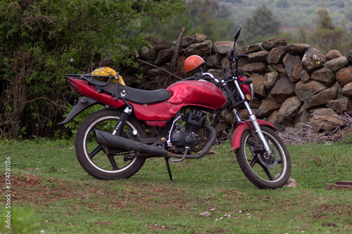 motocicleta de color guindo con negro de aspecto usado parada sobre un costado de un serco de piedras y in arbusto de color verde photo