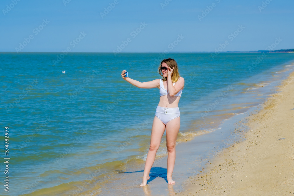 Beautiful young girl on the sea beach near the water posing and taking a selfie on a smartphone. Girl in a white swimsuit and sunglasses. Summer sunny day. Sea vacation concept. Copy space. Beautiful young girl on the sea beach near the water posing and taking a selfie on a smartphone. Girl in a white swimsuit and sunglasses. Summer sunny day. Sea vacation concept. Copy space.