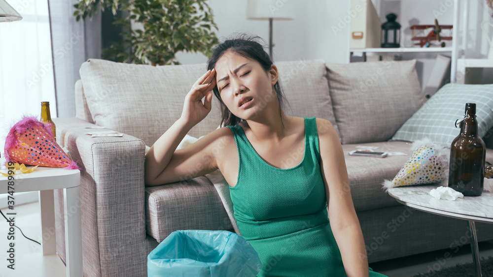 korean lady sitting by sofa suffering from hangover after party. sick young woman rubbing temples and vomiting in trash bin. real moments korean lady sitting by sofa suffering from hangover after party. sick young woman rubbing temples and vomiting in trash bin. real moments