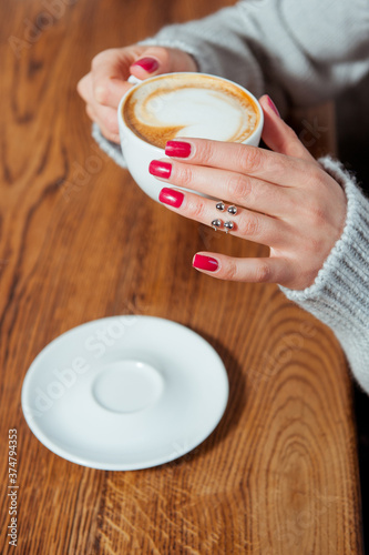 Young woman holding cup of coffee with latte art. She is wearing gray wool sweater and trendy silver ring. Morning in a cafe. photo