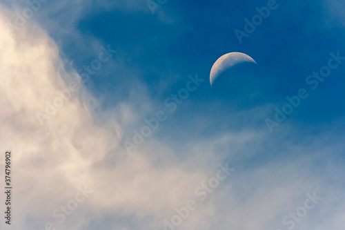 Crescent moon against a blue daytime sky with soft clouds around the moon. 