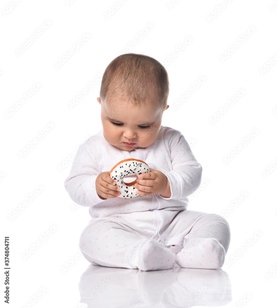 Focused infant baby toddler in white onepiece overall sits on the floor occupied with donut toy in hands Focused infant baby toddler in white onepiece overall sits on the floor occupied with donut toy in hands