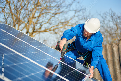 Man worker in blue suit and protective helmet installing solar photovoltaic panel system using screwdriver. Electrician mounting blue solar module on roof of modern house. Alternative energy concept.