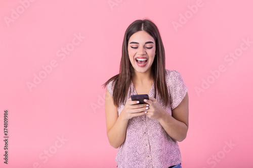 Adorable girl with mobile phone in hand expressing positive emotions. Attractive woman in pink shirt holding smartphone and texting on studio pink background
