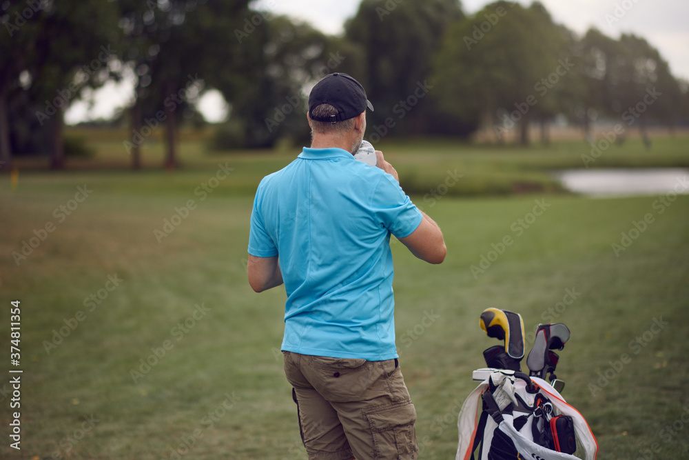 Golfer pausing for a drink on the golf course standing with his back to the camera alongside his golf bag and clubs looking down the fairway Golfer pausing for a drink on the golf course standing with his back to the camera alongside his golf bag and clubs looking down the fairway