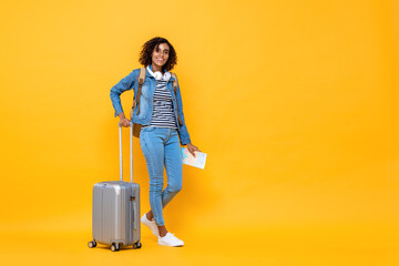 Full length travel portrait of smiling young African American woman backpacker standing and holding luggage on yellow studio background