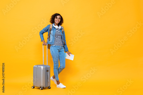 Full length travel portrait of smiling young African American woman backpacker standing and holding luggage on yellow studio background