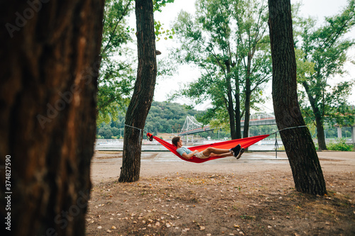 Valokuvatapetti Man relaxes lying in the park on a hammock hanging on a tree on the background of the river embankment and bridge