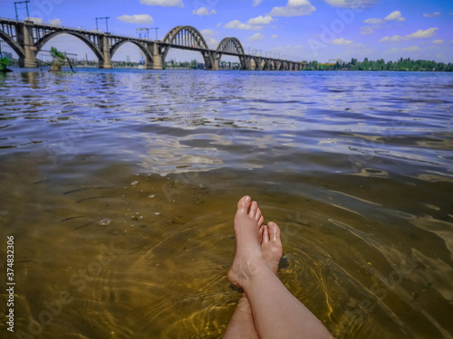 Wet female feet in the water against the background of a blurred landscape with the Merefa-Kherson bridge in Dnipro (Ukraine). Beautiful view from the beach on Monastyrskyi Island photo