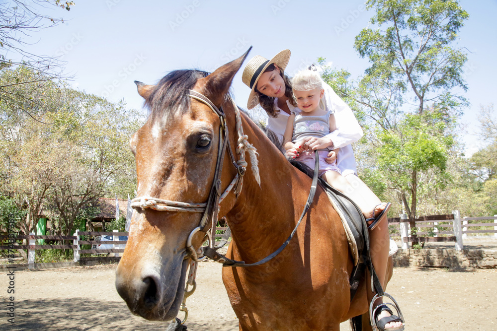 Fototapeta premium Beautiful young woman riding a horse with her little toddler girl, summer time outdoor activity