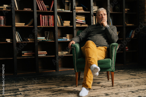 Worried bearded gray-haired elderly man thoughtfully holding hand with watch near chin, sitting at home on background of bookshelves in cozy room with an aristocratic interior, shooting from below.