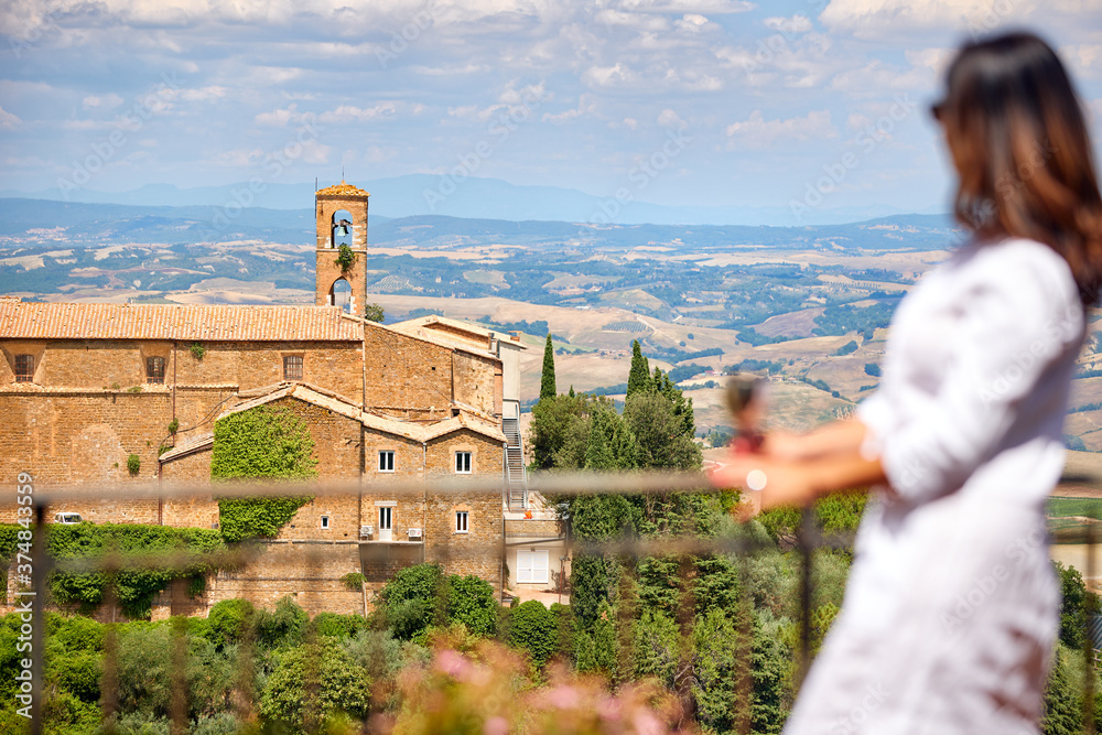 Holidays in Tuscany. Middle aged woman wearing a white dress watches the landscape of Tuscany from the terrace of a medieval house with a glass of wine in her hand. Montalcino, Italy.