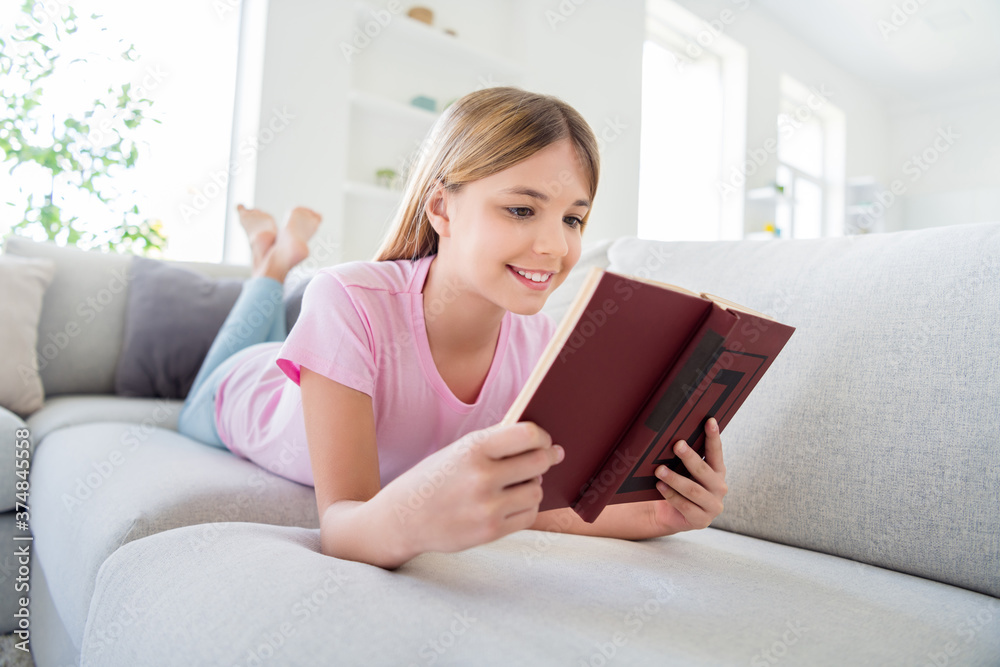 Full length photo of positive kid girl lying divan bare foot read book homeschool homework task wear t-shirt denim jeans in house indoors Full length photo of positive kid girl lying divan bare foot read book homeschool homework task wear t-shirt denim jeans in house indoors
