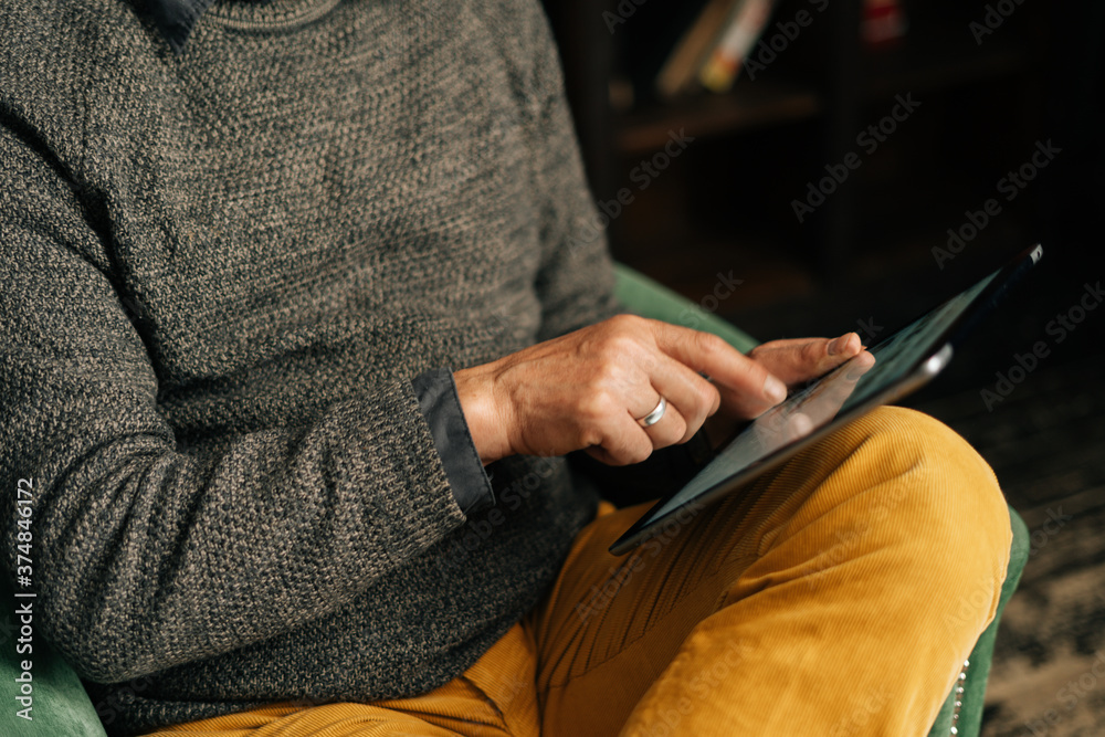 Close-up of hands of unrecognizable senior adult male browsing internet content using digital tablet, sitting at home in armchair in cozy living library room with an authentic aristocratic interior.